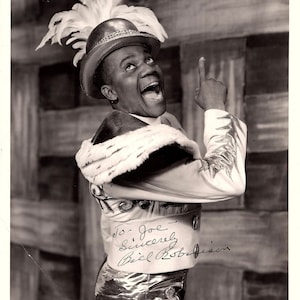 May include: Black and white photo of a man wearing a gold and white costume with a feathered hat. He is smiling and pointing upwards. The photo is signed "To Joe, Sincerely, Bill Robinson".