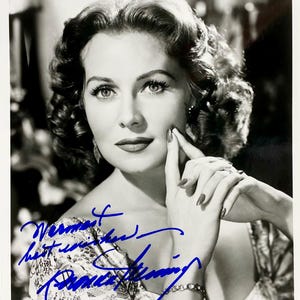 May include: Black and white photograph of a woman with dark, curly hair. She is wearing a patterned dress and has her hand on her chin. The photo is signed "Vermont best wishes Barbara Stanwyck".