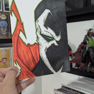 May include: Hand-drawn artwork of the comic book character Spawn, featuring a black and white face with green accents and a red cape. The artwork is held in a hand, with comic books and figurines in the background.