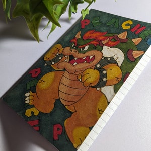 May include: A green and brown notebook with a cartoon illustration of Bowser from the Super Mario Bros. video game series. The notebook has a lined interior and the text "Peach" on the cover.