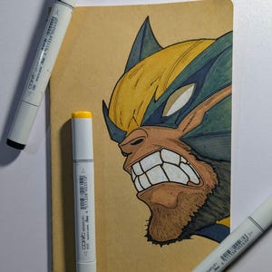 May include: A drawing of Wolverine with yellow and blue accents on a tan background. Three white Copic Sketch markers with black, yellow, and blue caps surround the drawing. The drawing features a close-up of Wolverine's face.