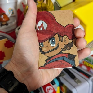 May include: A hand holds a small, brown notebook with a hand-drawn illustration of Mario from the Super Mario video game series. The illustration is in color and features Mario's signature red hat and overalls.
