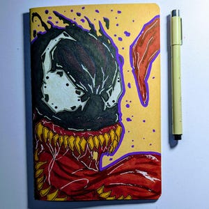 May include: A hand-drawn notebook featuring a Venom illustration. The artwork showcases Venom's face in black, white, and red, with yellow teeth and purple outlines. A yellow pen is placed next to the notebook.