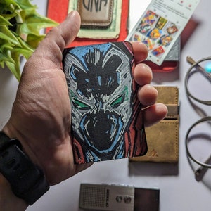 May include: A hand holding a black and white drawing of a comic book character's face. The character has a white mask with green eyes and a red cape. The drawing is on a small, rectangular piece of cardboard.