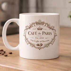 May include: White ceramic mug with a brown ornate design that reads "Caf&eacute; de Paris" with "Est. 1898" and "Fine Coffee & Chocolat". The mug has a curved handle and is on a wooden surface.