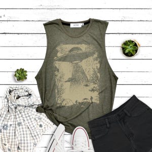 Puede incluir: Una camiseta verde sin mangas con un gráfico de estilo vintage de un OVNI flotando sobre un campamento. El gráfico está en tonos marrones y beige.