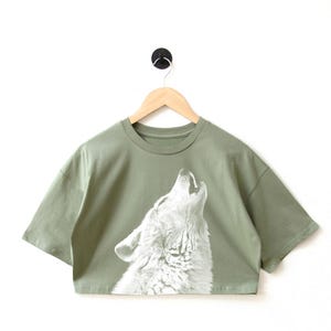 Pode incluir: Uma camiseta verde musgo cropped com uma estampa gráfica de um lobo branco uivando.