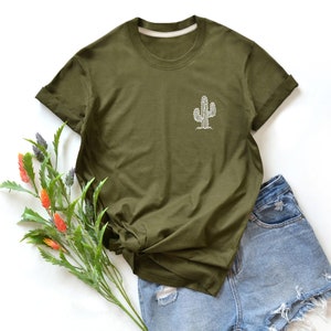 Puede incluir: Una camiseta verde de manga corta con un gráfico de cactus blanco en la parte delantera. La camiseta está sobre una superficie blanca con un par de pantalones cortos de mezclilla azul.