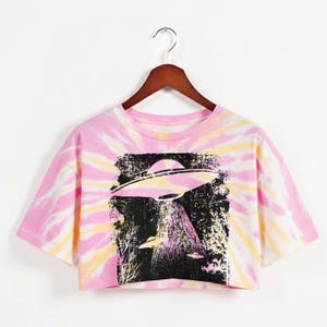 May include: Pink, yellow, and white tie-dye crop top with a black graphic of a UFO over a forest. The UFO graphic has a retro, vintage style. The crop top is hanging on a wooden hanger.