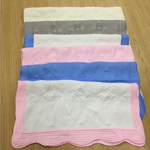 Puede incluir: Cinco mantas acolchadas de diferentes colores, apiladas unas sobre otras. Las mantas son blancas, grises, rosas, azules y blancas con un borde rosa. Las mantas tienen un borde festoneado y un patr&oacute;n decorativo.