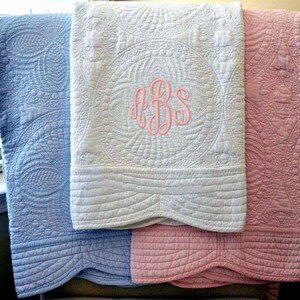May include: Three folded, quilted blankets in pastel colors: light blue, white, and pink. The white blanket features a pink monogram. Each blanket has a detailed, textured pattern. The blankets are likely for a baby or child.