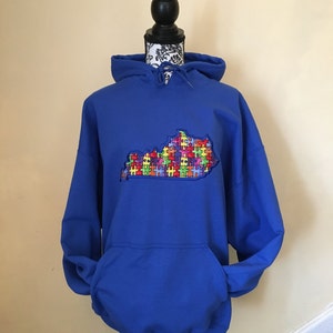 Op de afbeelding: Een koningsblauwe hoodie met een voorzak en een capuchon met trekkoord. De hoodie heeft een geborduurde omtrek van de staat Kentucky, gevuld met een kleurrijk puzzelstukontwerp, dat autismebewustzijn symboliseert.