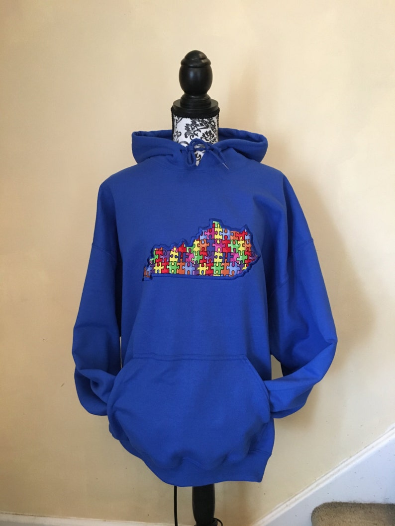 Op de afbeelding: Een koningsblauwe hoodie met een voorzak en een capuchon met trekkoord. De hoodie heeft een geborduurde omtrek van de staat Kentucky, gevuld met een kleurrijk puzzelstukontwerp, dat autismebewustzijn symboliseert.