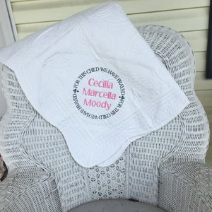May include: White quilted baby blanket with pink and black embroidery. The embroidery reads "For this child we have prayed + Cecilia Marcella Moody + For this child we have prayed".