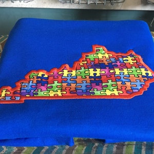 Op de afbeelding: Een opgevouwen blauwe deken met een geborduurde omtrek van de staat Kentucky. De omtrek van de staat is gevuld met kleurrijke puzzelstukjes, omrand met rood en geel.