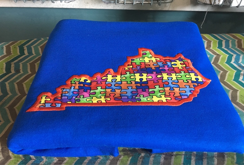 Op de afbeelding: Een opgevouwen blauwe deken met een geborduurde omtrek van de staat Kentucky. De omtrek van de staat is gevuld met kleurrijke puzzelstukjes, omrand met rood en geel.