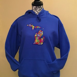 Op de afbeelding: Een koningsblauwe hoodie met een voorzak en een capuchon met trekkoord. De hoodie heeft een geborduurde omtrek van de staat Michigan, gevuld met een kleurrijk puzzelstukjesontwerp.