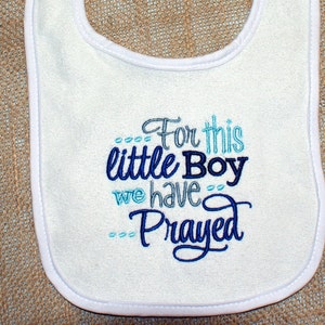 May include: White baby bib with blue and gray embroidery that reads "For this little Boy we have Prayed".