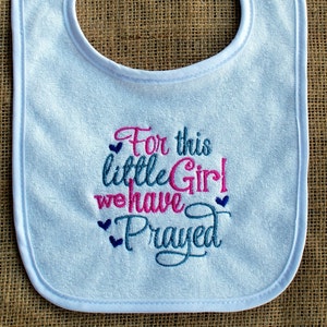 May include: A light blue baby bib with white trim. The bib features embroidered text in pink and blue that reads "For this little Girl we have Prayed". The bib is made of a soft, absorbent material.