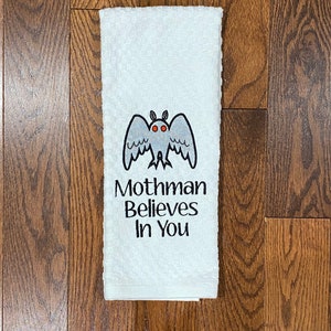 May include: White kitchen towel with an embroidered black and white mothman with red eyes. The text "Mothman Believes In You" is embroidered below the mothman.
