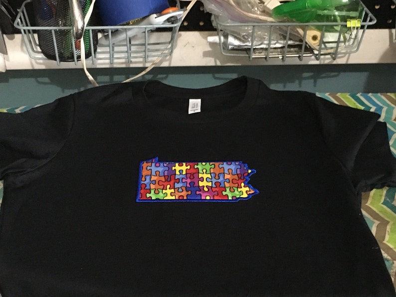 Op de afbeelding: Zwart T-shirt met een kleurrijk puzzelstukontwerp in de vorm van de staat Pennsylvania. De puzzelstukjes zijn in verschillende kleuren, omlijnd in blauw. Het shirt heeft een ronde hals en korte mouwen.