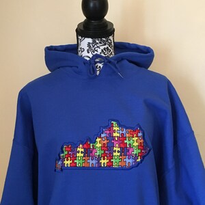 Op de afbeelding: Koningsblauwe hoodie met een geborduurde omtrek van Kentucky gevuld met een kleurrijk puzzelstukjesontwerp. De hoodie heeft een capuchon met trekkoord en een voorzak. Het ontwerp is een levendige mix van rood, geel, groen, blauw en paars.