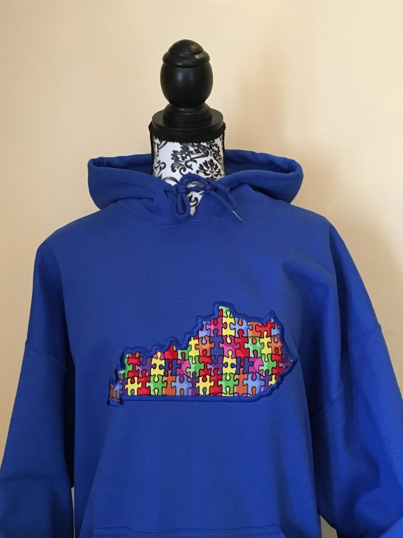 Op de afbeelding: Koningsblauwe hoodie met een geborduurde omtrek van Kentucky gevuld met een kleurrijk puzzelstukjesontwerp. De hoodie heeft een capuchon met trekkoord en een voorzak. Het ontwerp is een levendige mix van rood, geel, groen, blauw en paars.