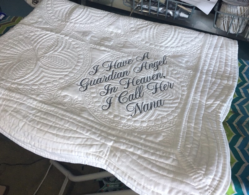 Puede incluir: Manta acolchada blanca con el texto bordado "Tengo un &aacute;ngel de la guarda en el cielo, la llamo Nana".