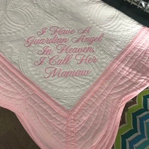 May include: White quilted blanket with pink trim and the embroidered text "I Have A Guardian Angel In Heaven, I Call Her Mamaw." The blanket features a detailed, patterned design and a scalloped edge.