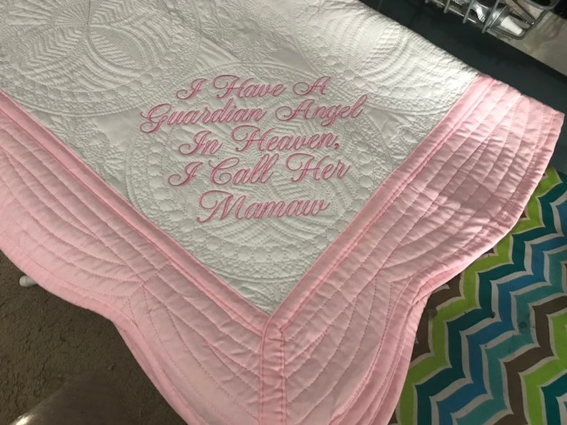 May include: White quilted blanket with pink trim and the embroidered text "I Have A Guardian Angel In Heaven, I Call Her Mamaw." The blanket features a detailed, patterned design and a scalloped edge.