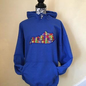 Op de afbeelding: Een koningsblauwe hoodie met een kangoeroezak en een capuchon met trekkoord. De voorkant heeft een geborduurde omtrek van de staat Kentucky, gevuld met kleurrijke puzzelstukjes. De hoodie wordt getoond op een zwarte mannequin.