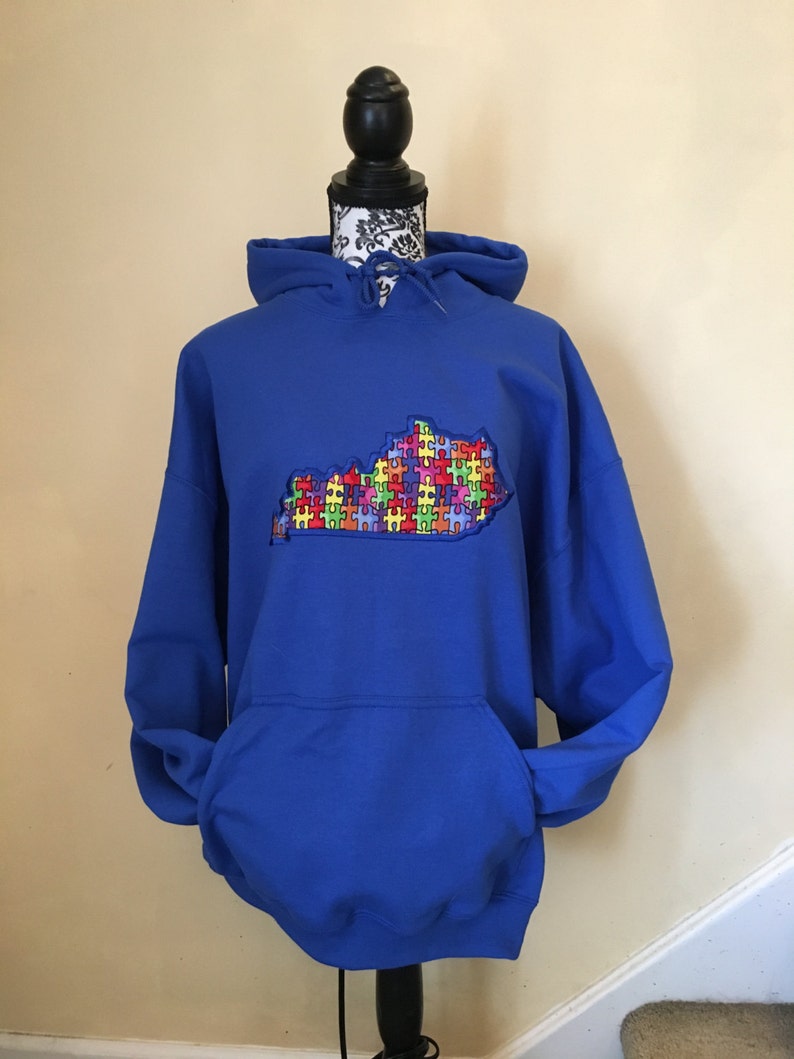 Op de afbeelding: Een koningsblauwe hoodie met een kangoeroezak en een capuchon met trekkoord. De voorkant heeft een geborduurde omtrek van de staat Kentucky, gevuld met kleurrijke puzzelstukjes. De hoodie wordt getoond op een zwarte mannequin.