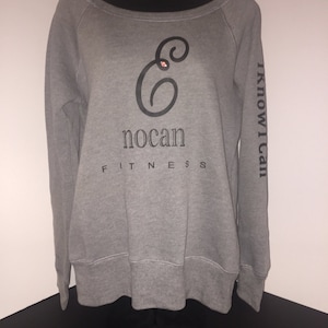 May include: A gray off-the-shoulder sweatshirt with the text "nocan FITNESS" and a stylized "e" logo on the front. The right sleeve has the text "I KNOW I CAN" printed vertically in black. The sweatshirt is displayed on a black mannequin.