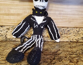 May include: A handmade crochet doll of Jack Skellington from The Nightmare Before Christmas. The doll has a white head with black eyes and a stitched mouth. It wears a black suit with white stripes and a white shirt. The doll is sitting.