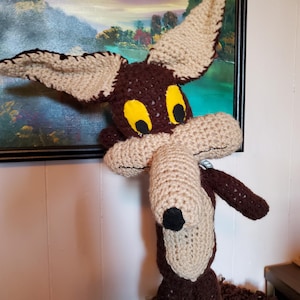 May include: A handmade crochet plush toy of a cartoon coyote. The coyote has brown and tan yarn with yellow eyes and a black nose. The coyote is standing in front of a painting.