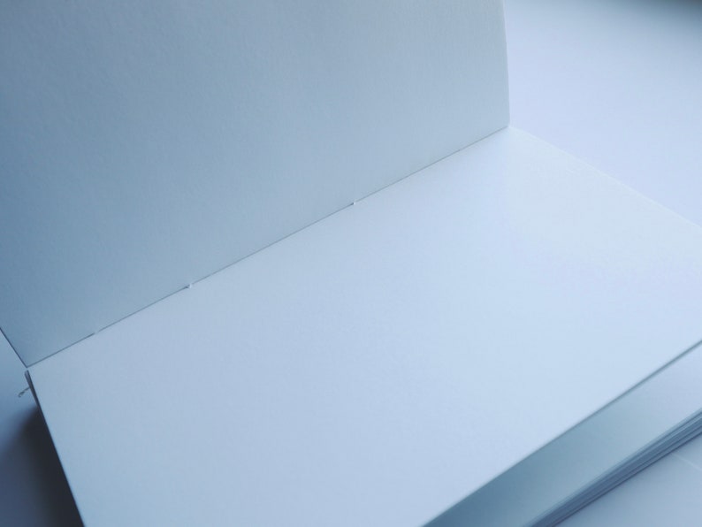 May include: A blank, white notebook with a spine and a few pages visible. The notebook is open and lying flat on a white surface.