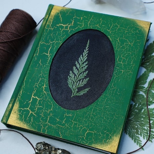 May include: A green journal with a distressed, cracked finish and gold accents. A circular cutout on the front features a pressed fern on a black background.