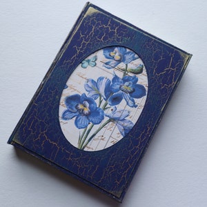 May include: A blue, distressed, wooden box with a gold border. The box has a circular cutout with a floral design featuring blue flowers and a butterfly.