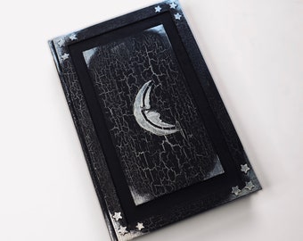 Puede incluir: Un diario negro con una luna creciente plateada y estrellas en la portada. La portada tiene un aspecto de pintura desgastada y agrietada.
