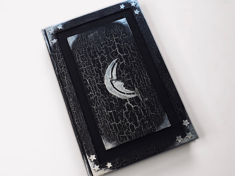 Puede incluir: Un diario negro con una luna creciente plateada y estrellas en la portada. La portada tiene un aspecto de pintura desgastada y agrietada.