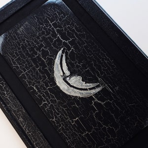 Puede incluir: Una caja de madera negra con un dise&ntilde;o de luna creciente plateada. La caja tiene un aspecto desgastado y envejecido con un acabado de pintura agrietada.