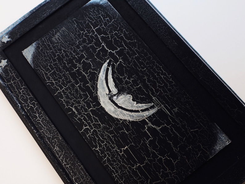 Puede incluir: Una caja de madera negra con un dise&ntilde;o de luna creciente plateada. La caja tiene un aspecto desgastado y envejecido con un acabado de pintura agrietada.