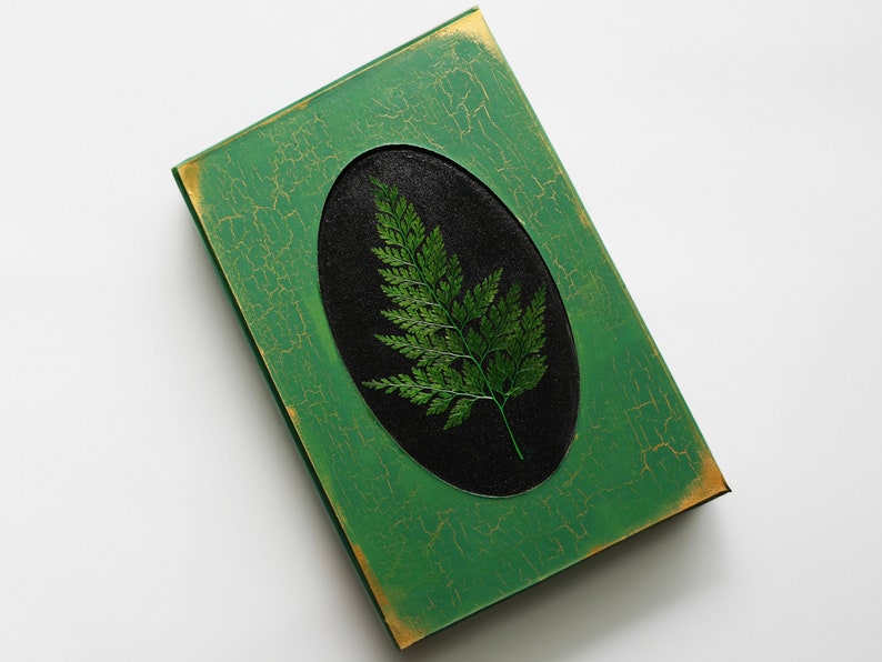 May include: A green wooden box with a distressed finish and a gold oval frame. Inside the frame is a pressed fern leaf on a black background.
