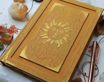 Puede incluir: Un diario encuadernado en cuero amarillo con un diseño de sol dorado en la portada. El diario tiene un aspecto desgastado y vintage.