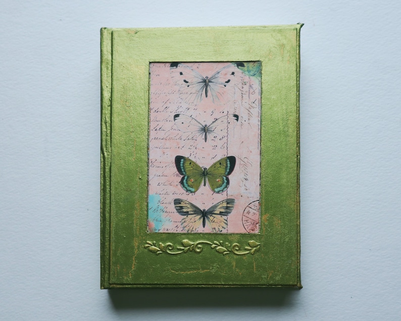 May include: A green journal with a gold frame and a pink and white butterfly design. The journal has a vintage look and feel.