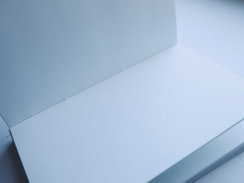 Puede incluir: Un cuaderno blanco en blanco con una encuadernaci&oacute;n y algunas p&aacute;ginas visibles. El cuaderno est&aacute; abierto y acostado plano sobre una superficie blanca.