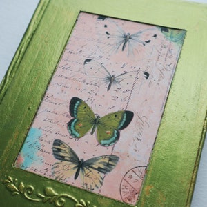 May include: A green framed picture with a pink background featuring a vintage postcard with handwritten text and four butterflies. The butterflies are green, white, and brown with black markings.