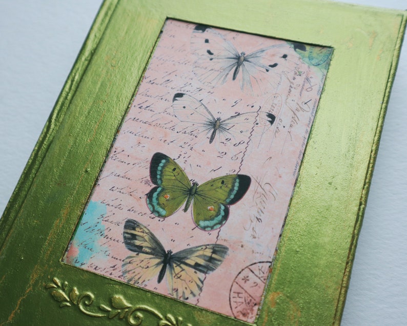 May include: A green framed picture with a pink background featuring a vintage postcard with handwritten text and four butterflies. The butterflies are green, white, and brown with black markings.