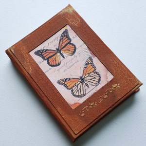 May include: A brown leather-bound journal with a distressed finish. The cover features a vintage-style illustration of two butterflies with text that reads "The Royal Institution 18... from the... Imparted respectfully by Mr. Griffith London".