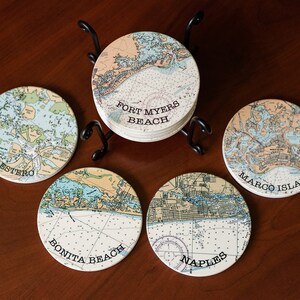 May include: Set of six round ceramic coasters featuring nautical maps of different Florida locations. The coasters are arranged in a circle on a wooden surface with a metal stand in the center. The coasters feature the names "Estero", "Bonita Beach", "Naples", "Marco Island", and "Fort Myers Beach".